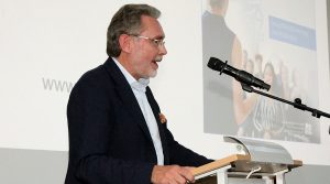 Dr. Torsten Tommpert spricht am Rednerpult mit Mikrofon und Pr&auml;sentation im Hintergrund.